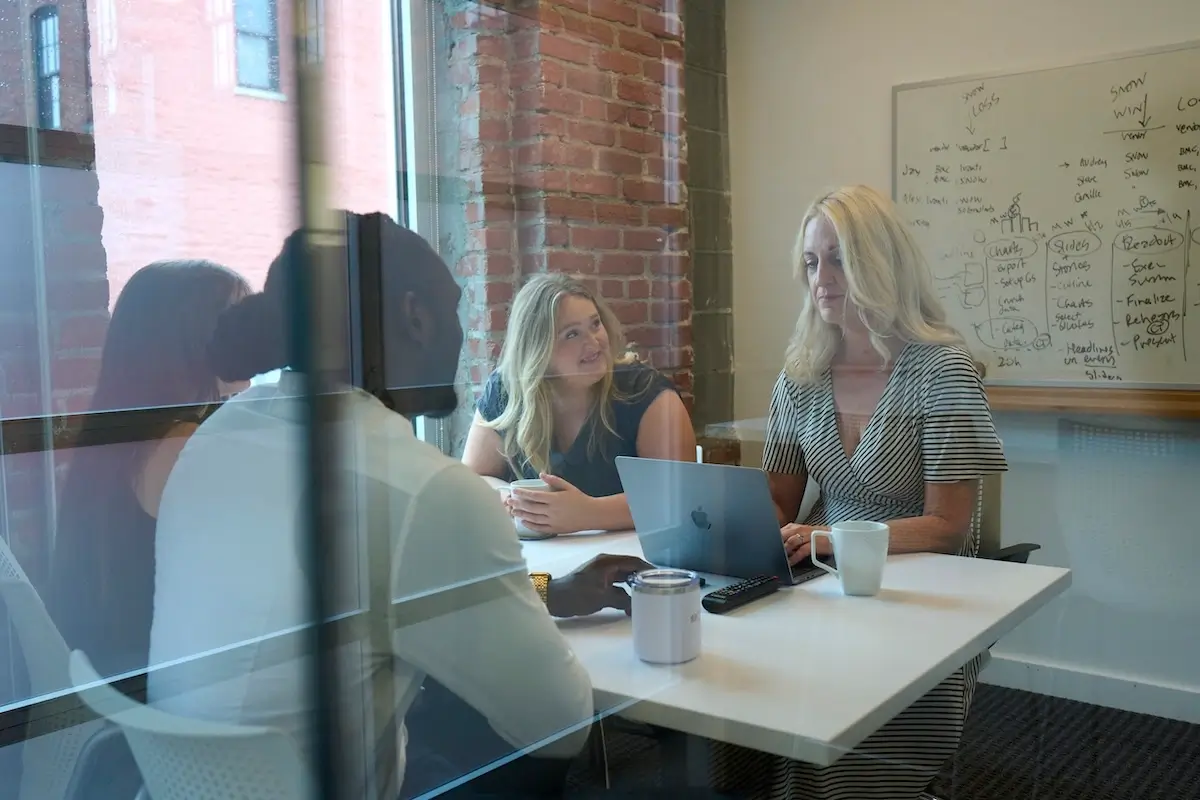 Four people sit around a white table in a glass-walled meeting room, discussing while one woman works on a laptop with notes on a whiteboard behind.