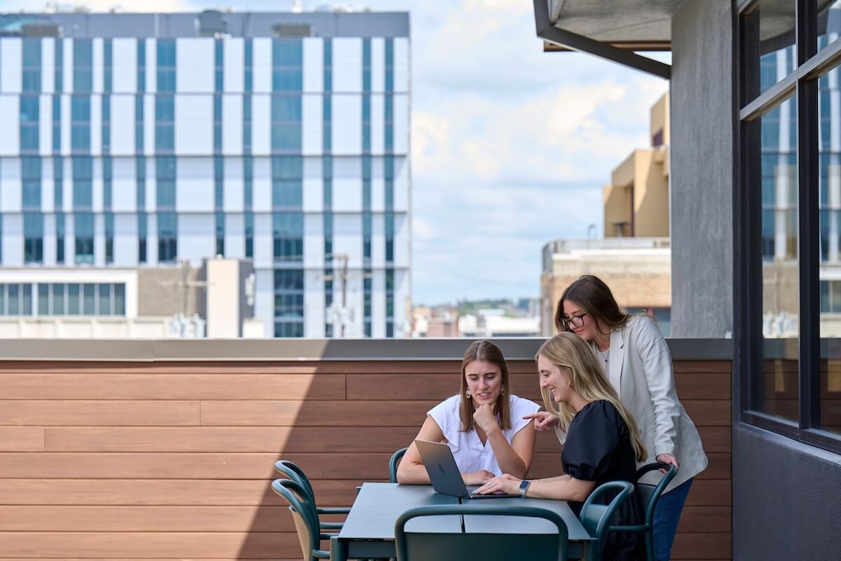 Three women sit and stand around a laptop at a patio table, smiling and discussing work with a city building in the background.